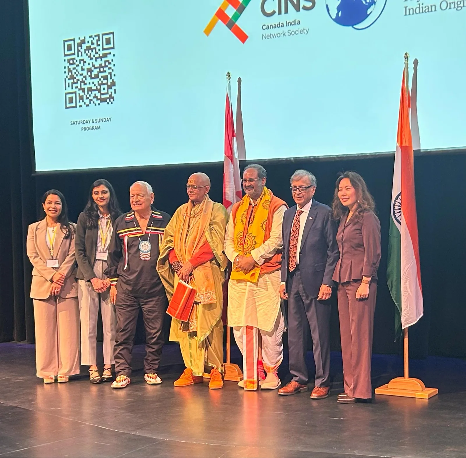 Seven keynote speakers stand together on a stage between Canadian and Indian flags, including an elder in a ribbon shirt and beaded medallion and two men in saffron robes and shawls, beneath a Canada India Network Society screen.