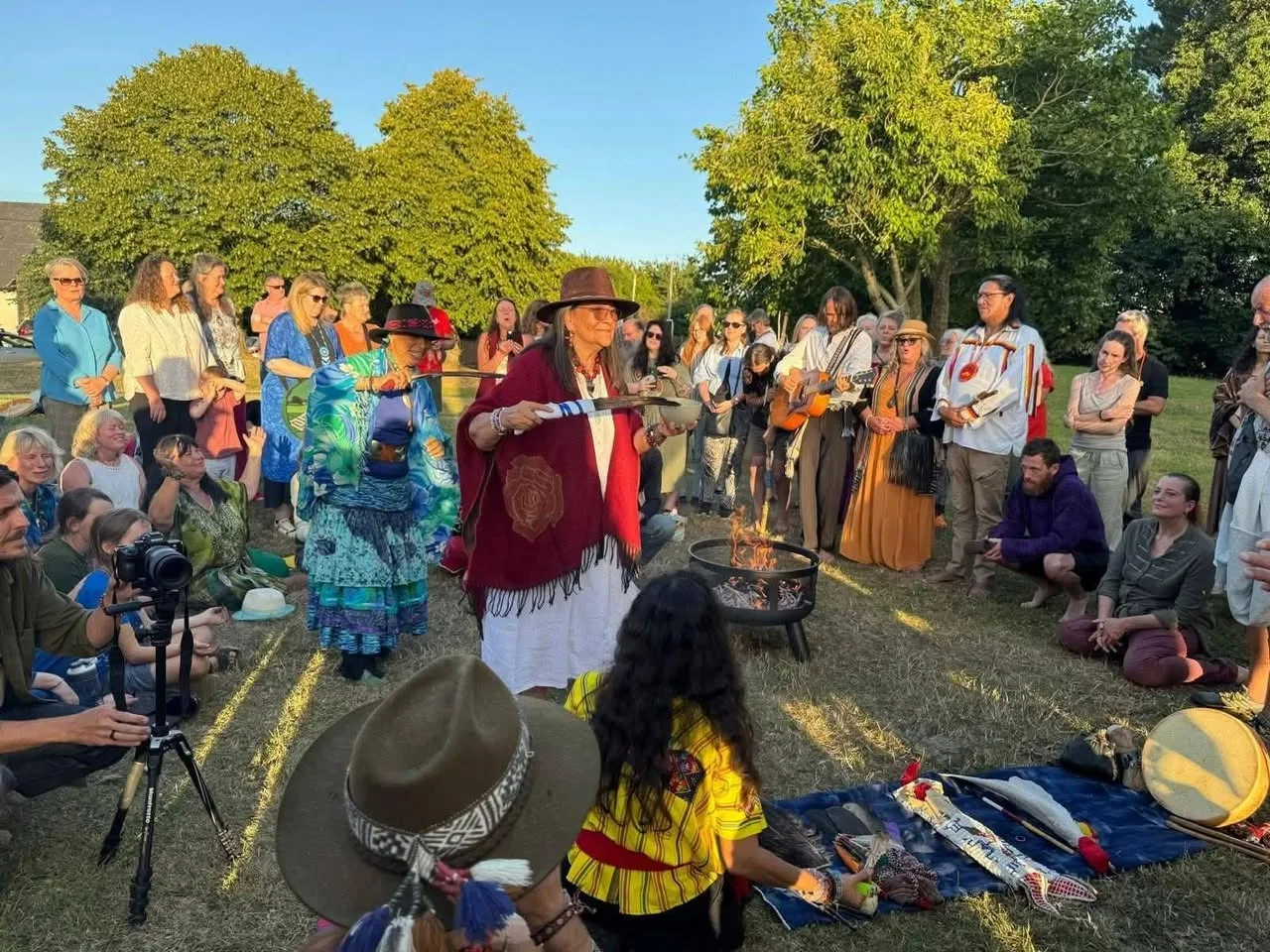 A large circle of people gathers on grass at sunset around a fire in a metal bowl, as women in shawls and hats hold feathers and a bowl in ceremony, with a guitarist and drum nearby.