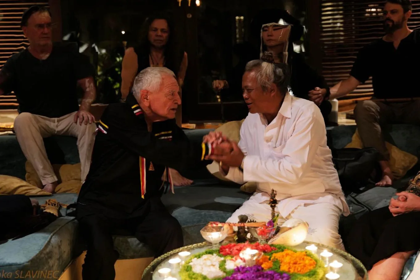 An elderly white-haired man in a black ribbon shirt clasps hands with a smiling Indonesian priest in white, beside a tray of flowers and candles, as others sit watching.