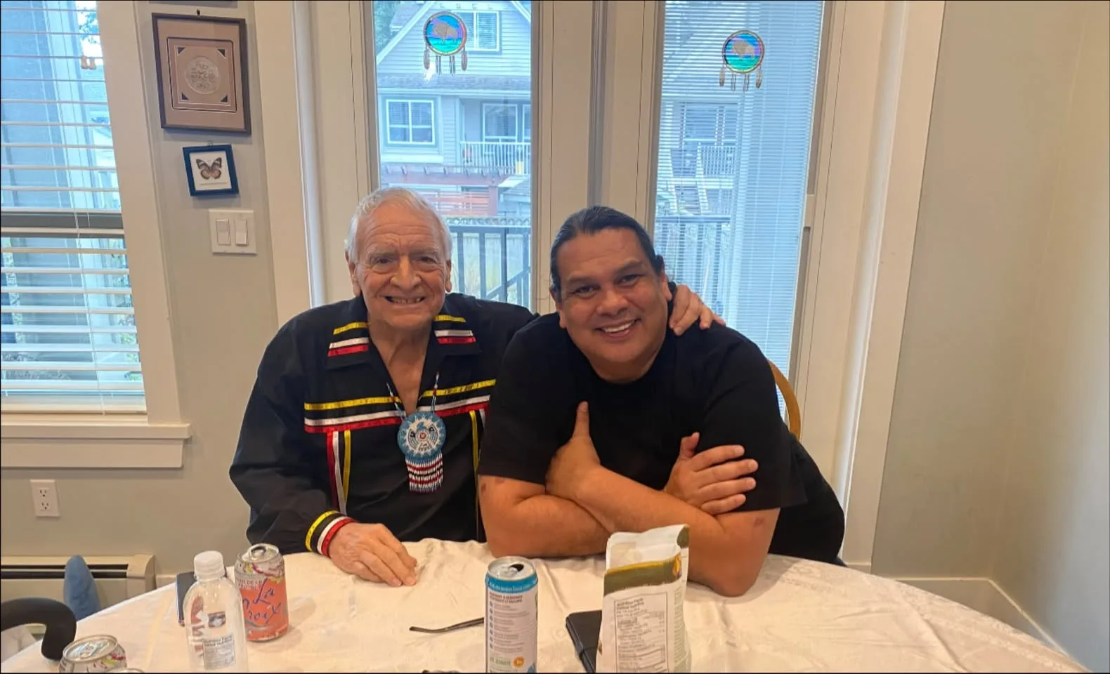 An elderly man with white hair, wearing a black ribbon shirt and beaded medallion, sits smiling at a kitchen table beside a younger man in a black T-shirt with his arms crossed.