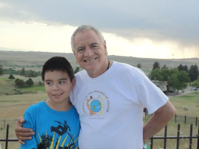 A smiling gray-haired man in a white Center for the Healing of Racism T-shirt hugs a boy in a blue shirt beside an iron fence, with grassy hills and a stormy sky behind.