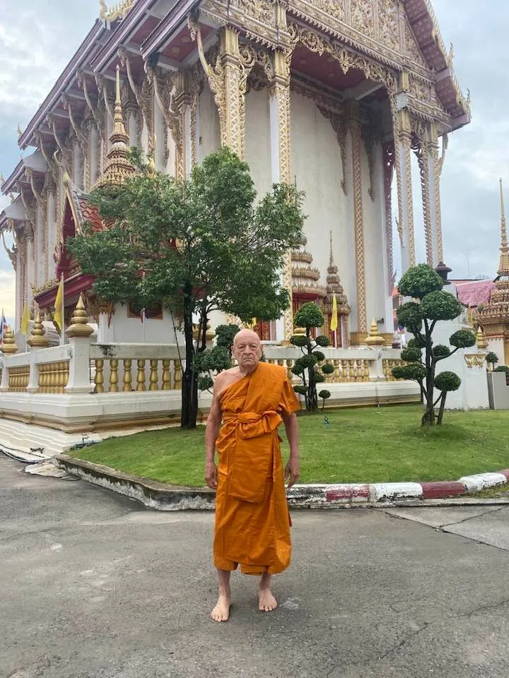 An elderly man with a shaved head, barefoot in saffron monk's robes, stands before an ornate white and gold Thai Buddhist temple.