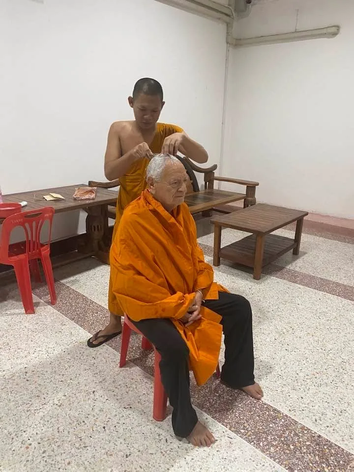 A young monk in saffron robes shaves the white hair of an elderly man seated on a red plastic chair, an orange cloth draped over his shoulders.