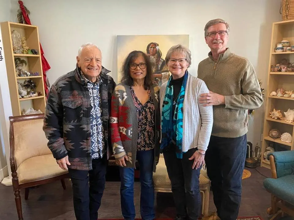 Four smiling people stand together in a room with shelves of shells and crafts, a painting of an Indigenous figure on the wall behind them; likely Phil, Suthida, Ruth and Joel.