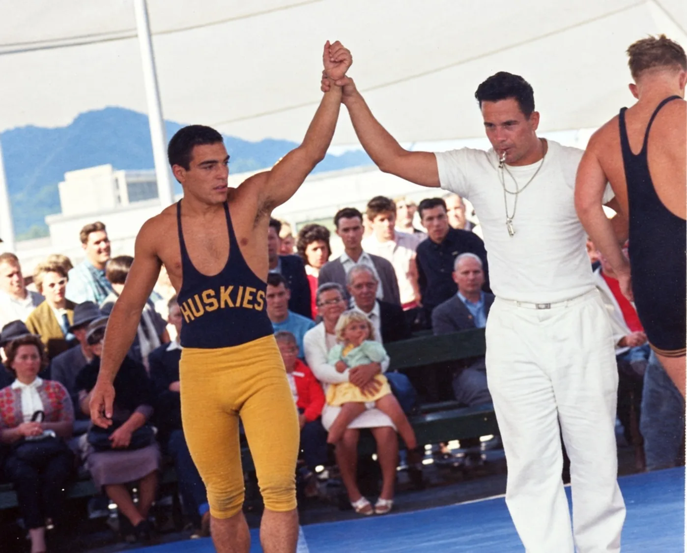 A young wrestler in a navy Huskies singlet and gold tights has his arm raised in victory by a referee in white with a whistle, as spectators watch from benches outdoors.