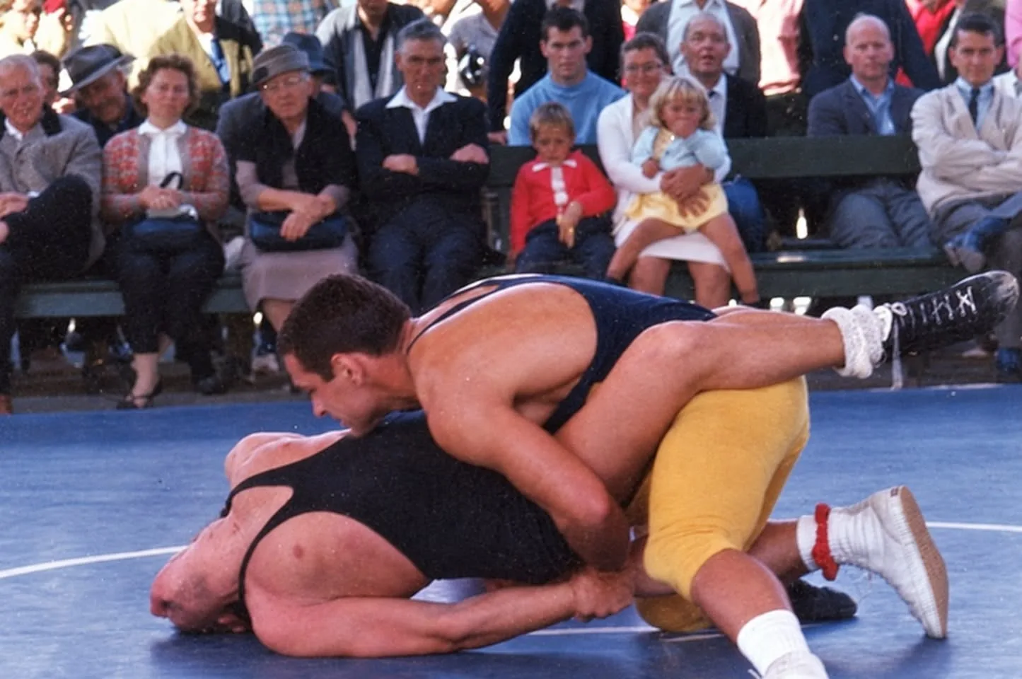 A wrestler in a navy singlet and gold tights pins an opponent in black to a blue mat, while a crowd of adults and small children watches without expression.